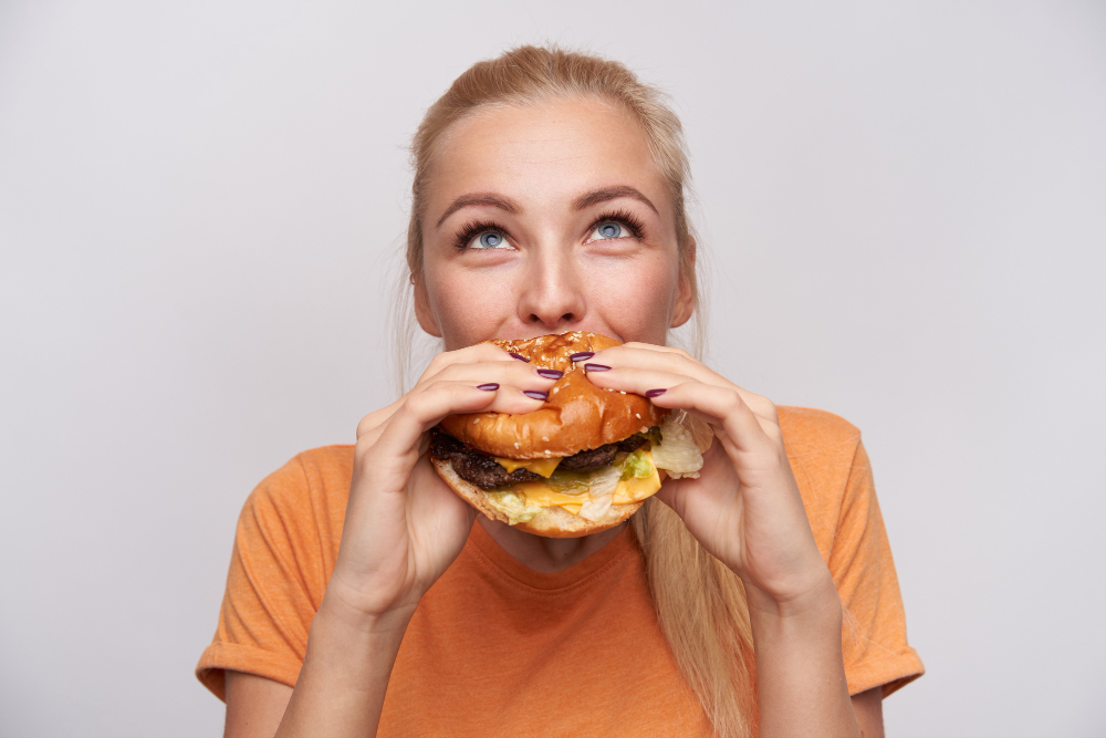 Portrait Pleased Young Lovely Blonde Woman With Casual Hairstyle Eating Fresh Hamburger With Great Appetite Looking Cheerfully Upwards Posing White Background