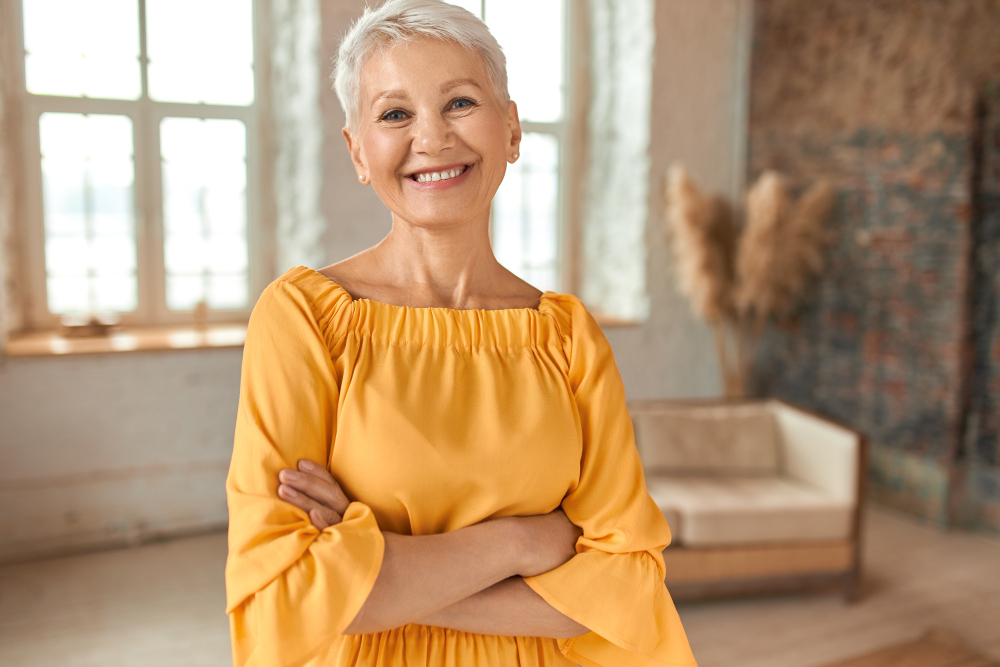 Attractive Confident Mature Blonde Woman Wearing Yellow Dress Crossing Arms Chest Smiling Happily Camera Posing Her Newly Renovated Apartment With Sofa Windows Background